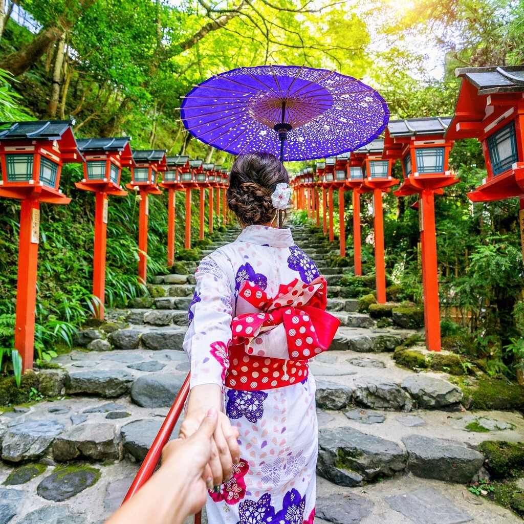 Nő hagyományos japán ruhában, lila esernyőt tartva, erdőn keresztül sétál egy szentély felé