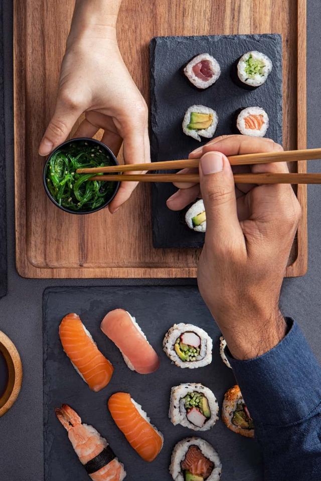 Válogatott sushi, beleértve a nigirit, sashimit és tekercseket, amelyeket pálcikákkal osztanak meg és esznek.